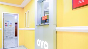 Un pasillo con paredes amarillas, una recepción identificada con 'OYO' y un letrero que indica servicio las 24 horas.