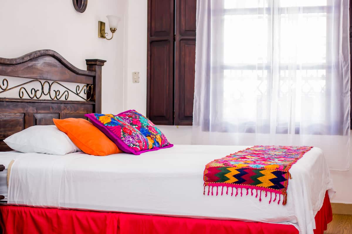 A neatly made bed with a colorful quilt and pillows, a wooden headboard with a decorative sign, and a window with sheer curtains.