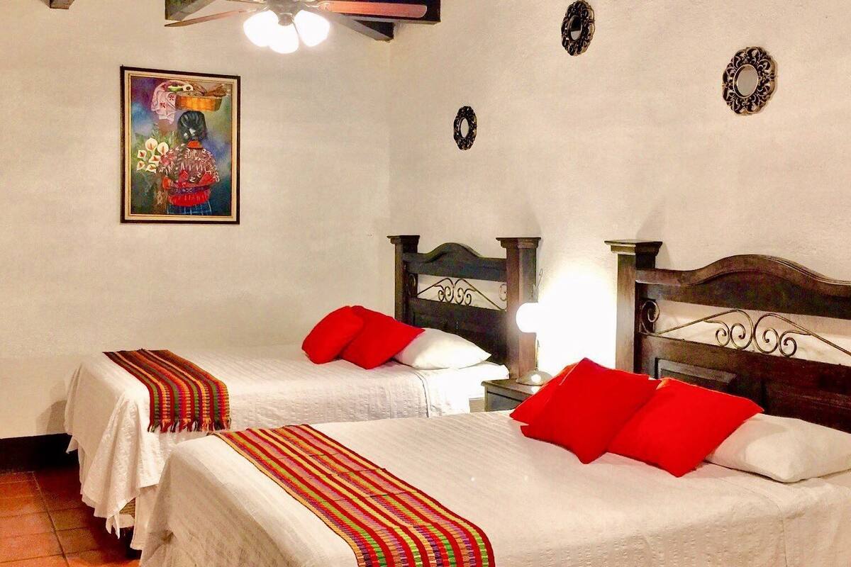 A hotel room with two beds, each with a red pillow, a white bedspread, and a colorful striped blanket. There is a ceiling fan, a wall clock, and a framed picture on the wall.