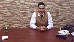 Un homme est assis à un bureau dans une pièce dont un mur est décoré d’un motif en pierre et sur laquelle est affiché un panneau indiquant « Villa Real ».