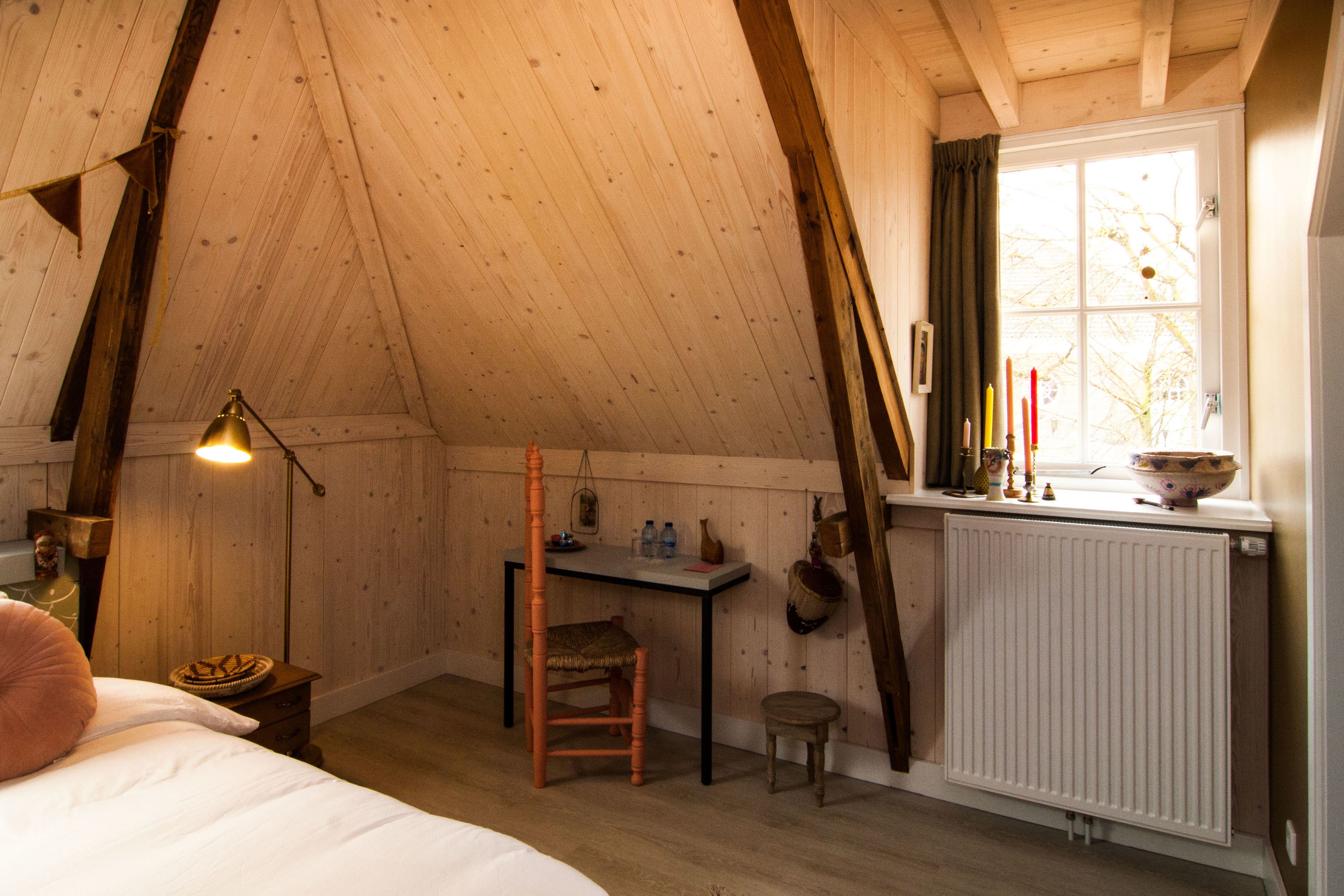 Ein gemütliches Schlafzimmer im Dachgeschoss mit schrägen Wänden, einem Holztisch, einem Stuhl, einem kleinen Hocker und einer Heizung.