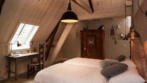 Quarto com teto inclinado, escrivaninha de madeira, cadeira, cama com roupa de cama branca e uma janela grande.