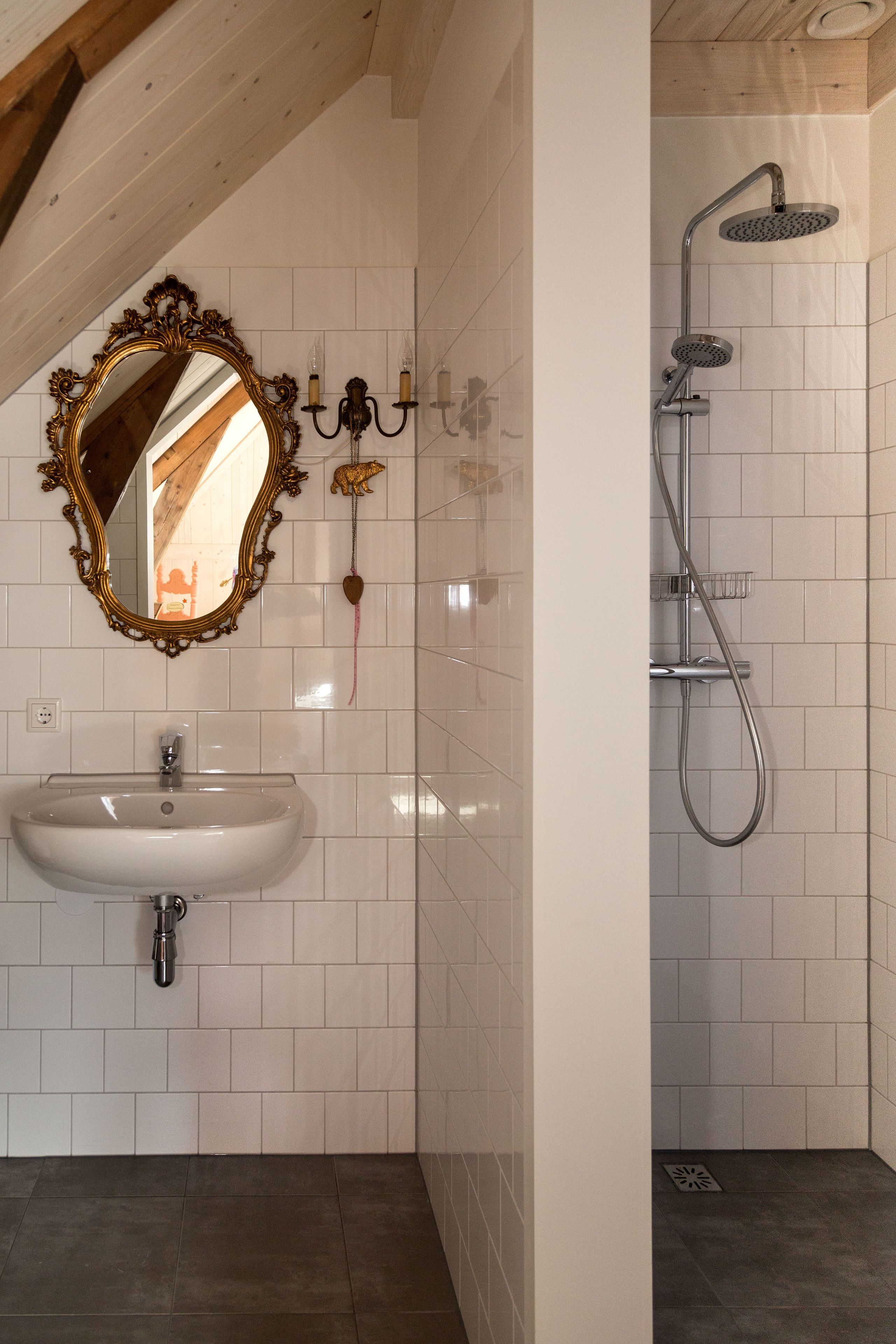 A bathroom with a white tiled wall, a white sink, a mirror with an ornate frame, a shower with a handheld showerhead, and a towel rack.