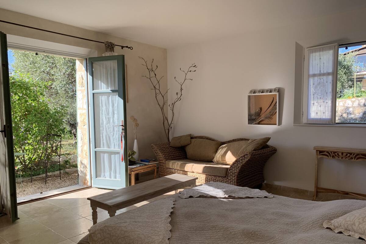 A cozy room with a bed, a bench, a sofa, and a table. There's a window with a view of greenery outside.