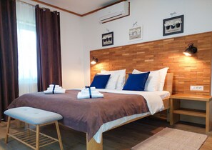 A neatly arranged hotel room with a wooden headboard, a bed with white and blue bedding, a bedside table, a bench, and a large window with curtains. - Guesthouse Lower Lakes (Plitvicka Jezera)
