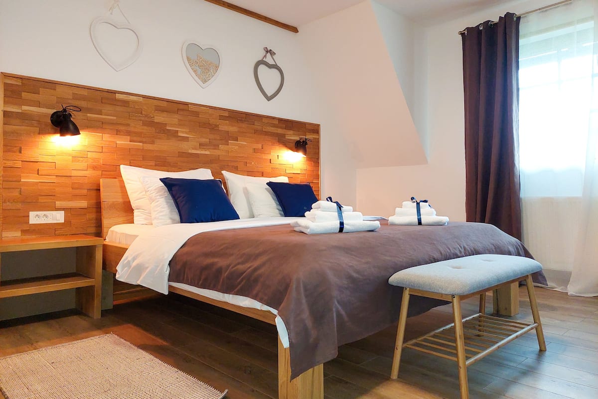 A modern bedroom with a wooden headboard, a bed with white and blue bedding, a bedside table, a bench, and a wall with heart-shaped decorations.