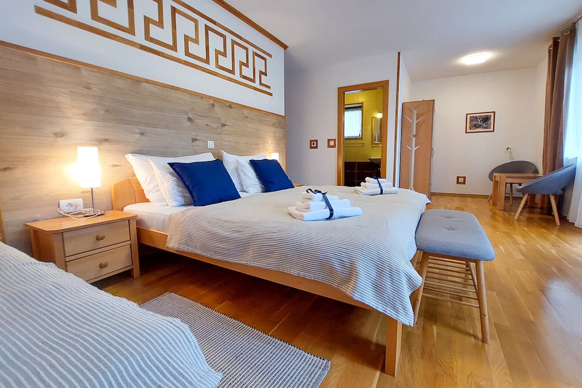 A modern hotel room with a wooden headboard, a large bed with blue and white bedding, a bedside table with a lamp, a bench, and a view of the hallway.
