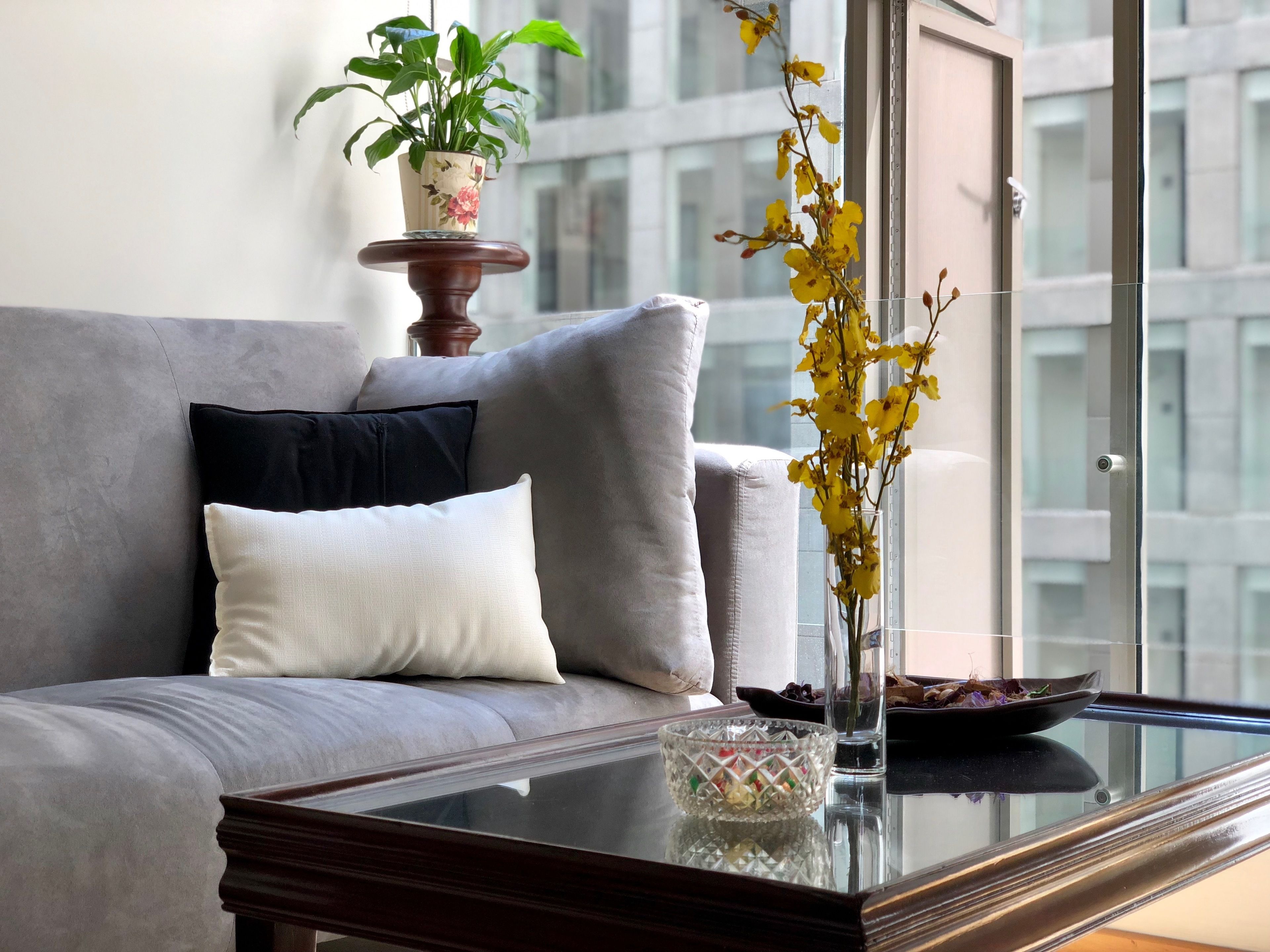 A modern living room with a grey sofa, a white cushion, a wooden side table, a glass vase with yellow flowers, and a potted plant on a wooden stand.