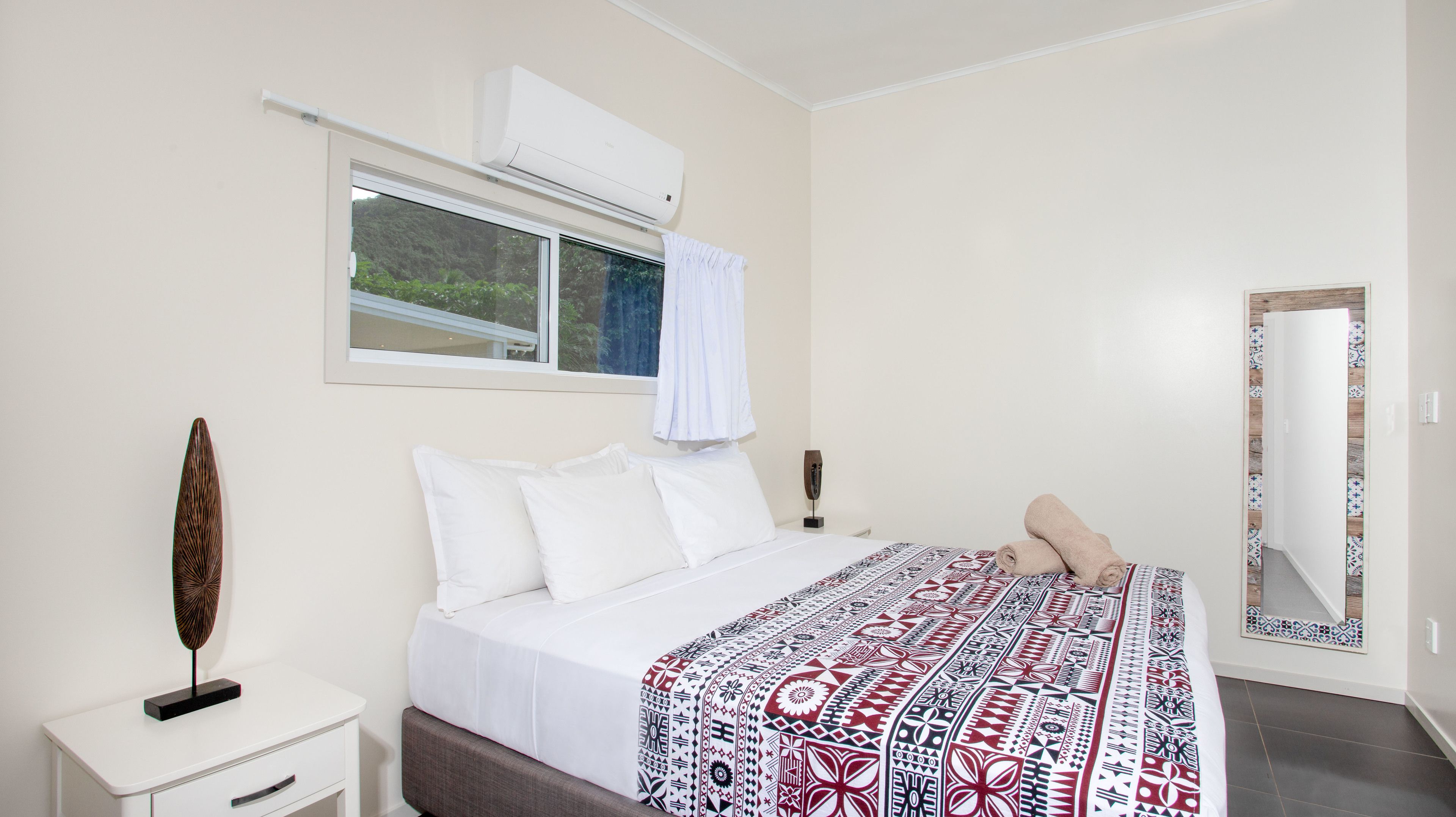 A bedroom with a bed, a bedside table, a window with a curtain, an air conditioning unit, and a mirror on the wall.