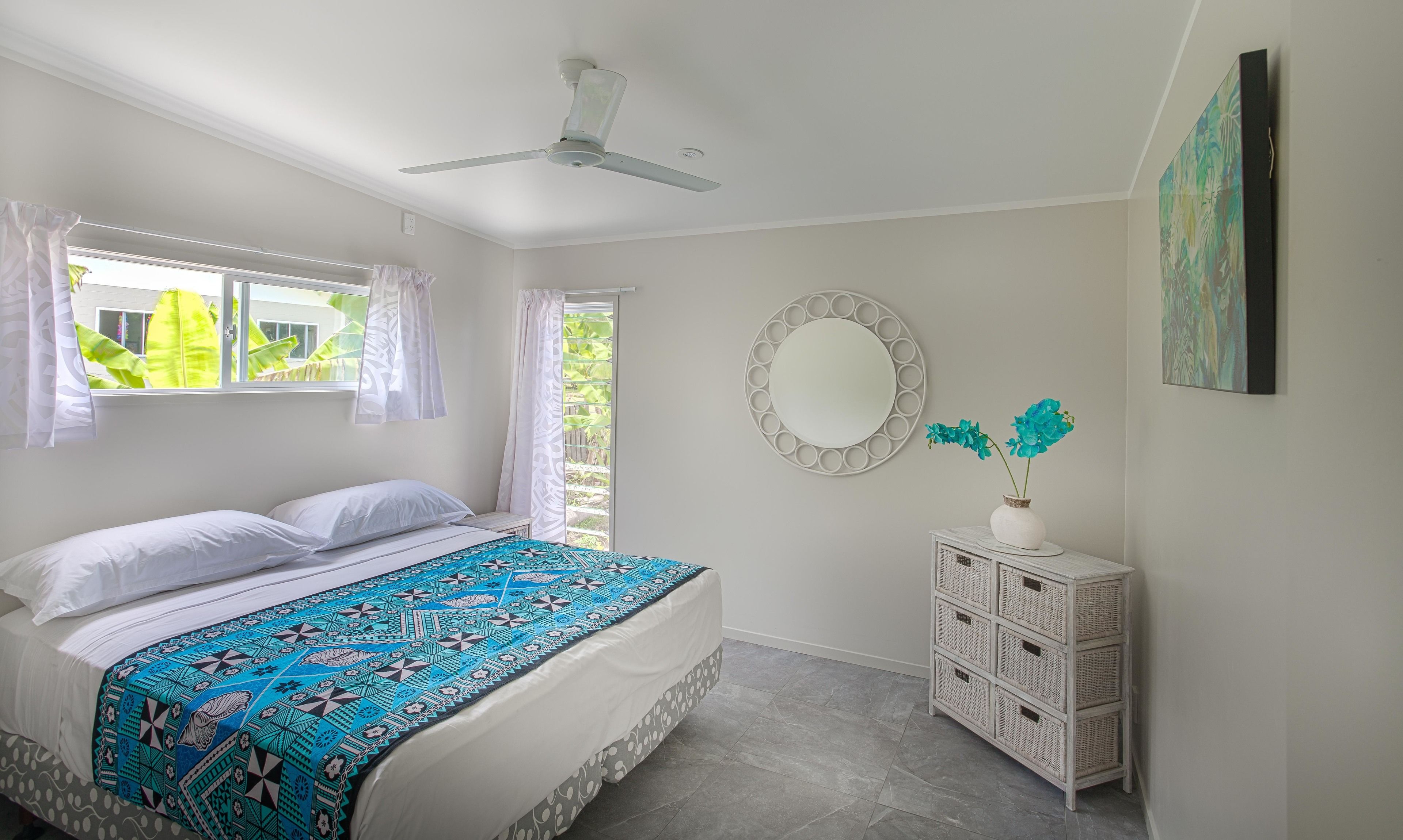 Ein Schlafzimmer mit einem Bett, Deckenventilator, Fenster mit Vorhängen, einem Spiegel und einer Kommode mit Schubladen.