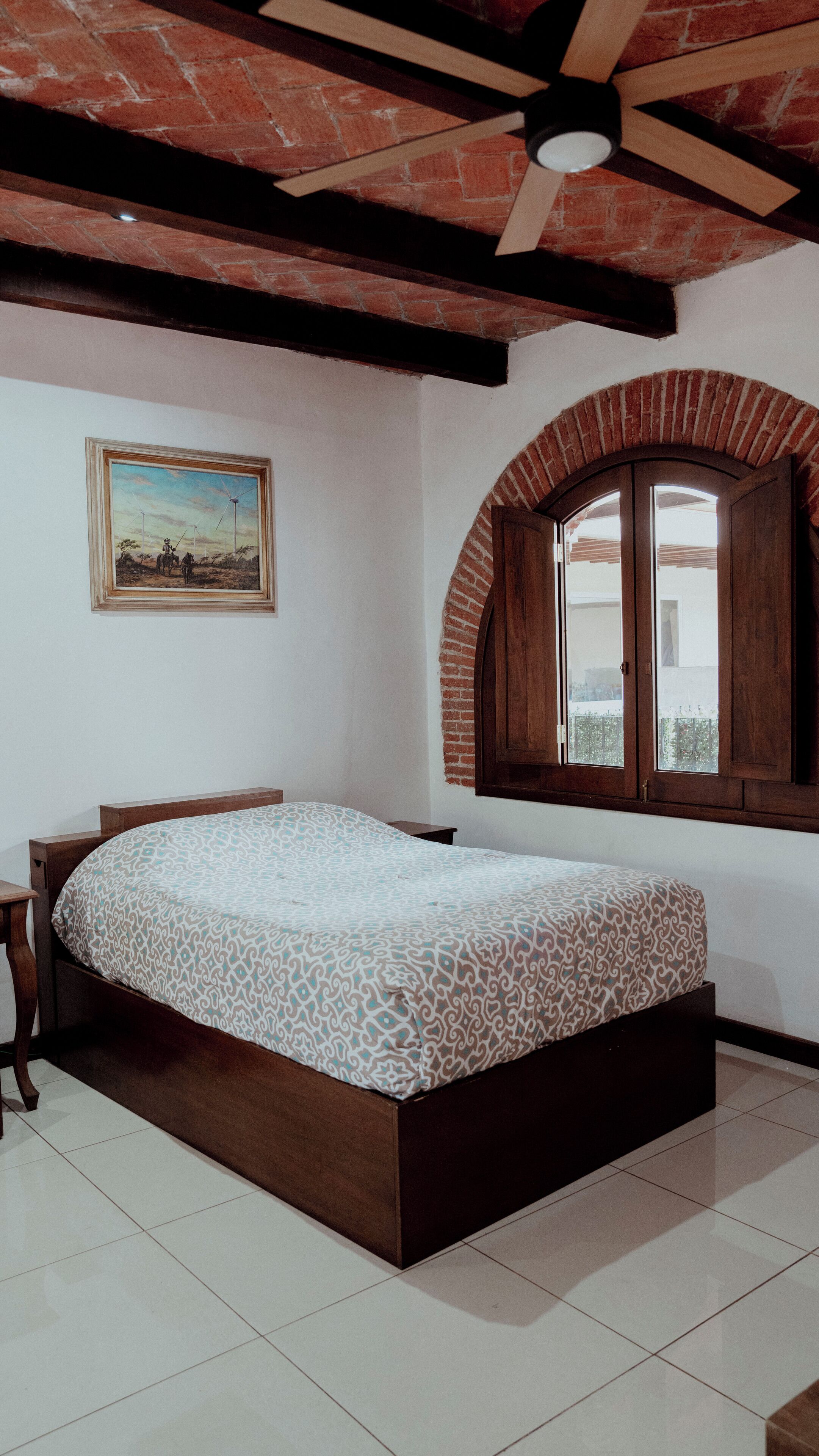 Un dormitorio con cama, cabecera de madera, un cuadro en la pared, una ventana con arco de ladrillo y un ventilador de techo.