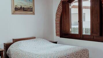 Un dormitorio con cama, cabecera de madera, un cuadro en la pared, una ventana con arco de ladrillo y un ventilador de techo.