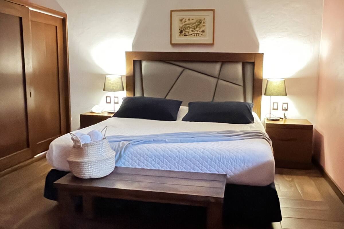 Ein ordentlich bezogenes Bett mit Kissen, ein Nachttisch mit Lampe und ein gerahmtes Bild an der Wand.