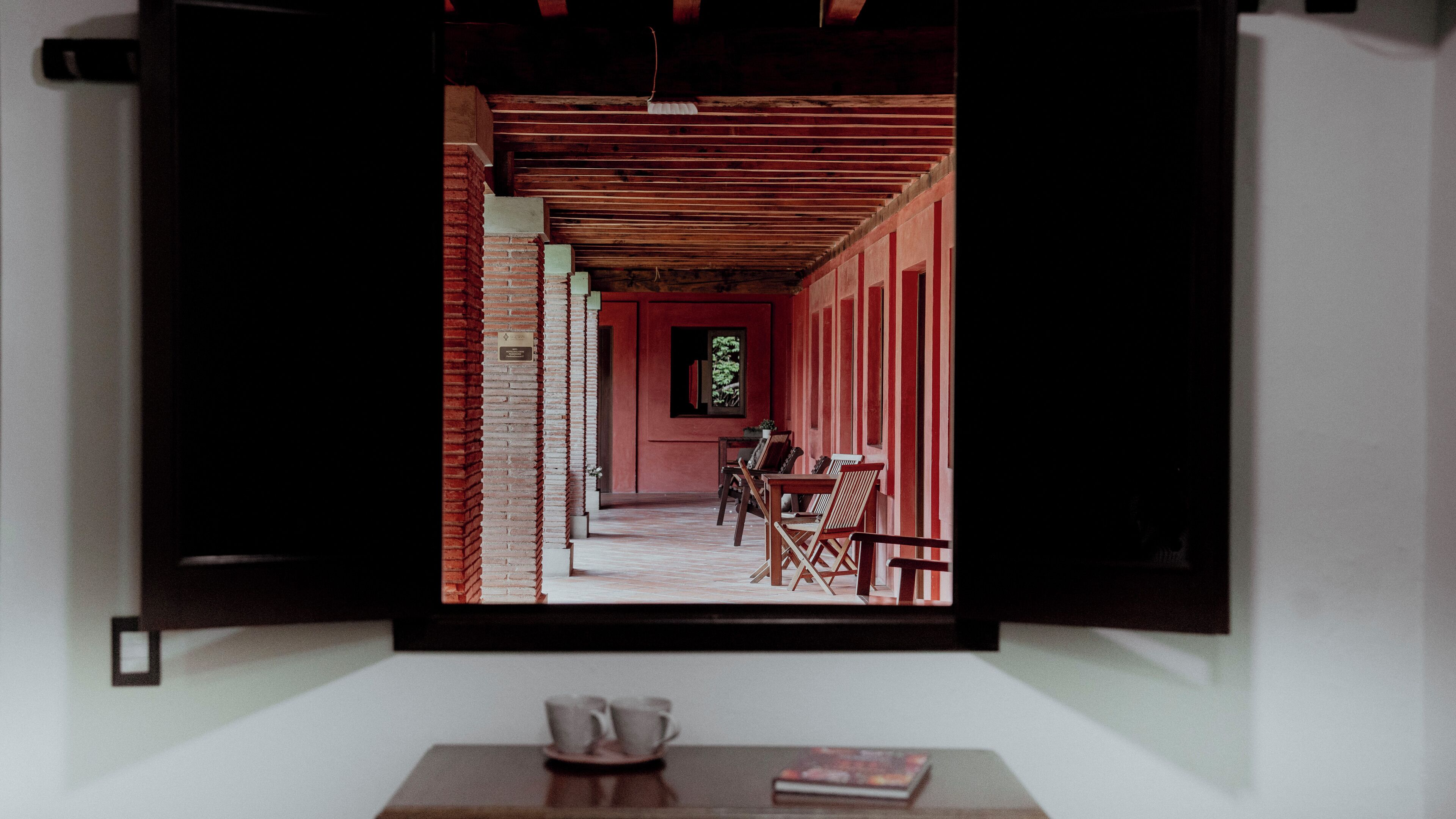 Una habitación con televisión mostrando un pasillo con pared roja, techo de madera y una ventana. Hay dos sillas y una mesa con tazas y un libro.
