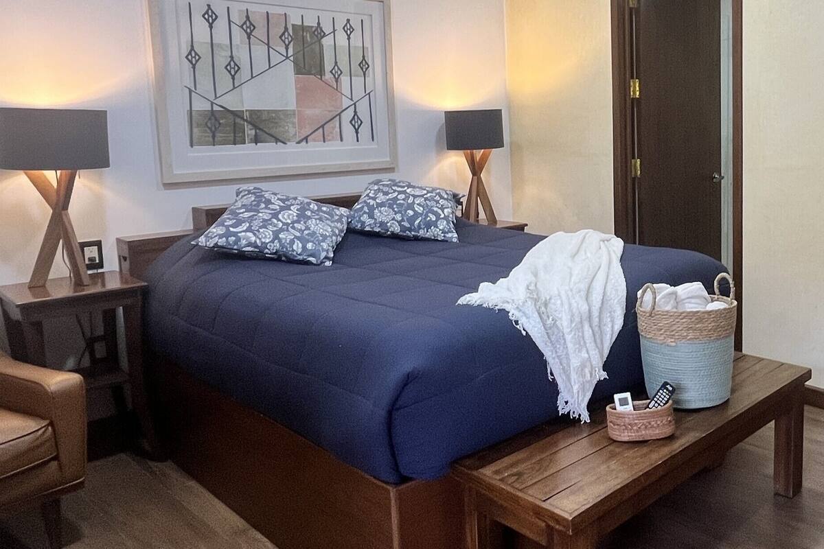 Ein ordentlich eingerichtetes Schlafzimmer mit einem großen Bett, zwei Nachttischlampen, einem Sessel, einem Nachttisch und einem gerahmten Kunstwerk an der Wand.