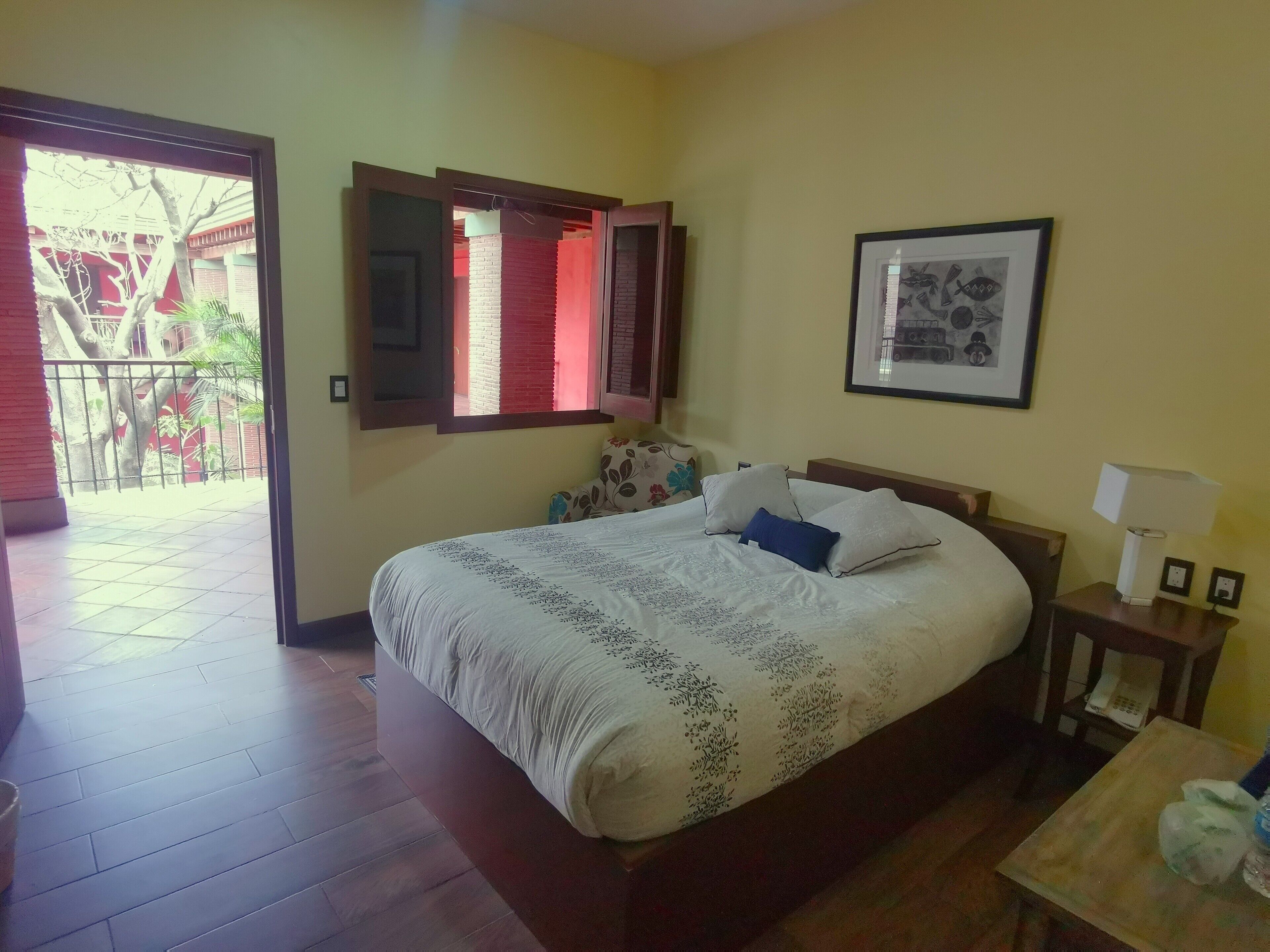 A hotel room with a bed, a bedside table with a lamp, a framed picture on the wall, and a view of the outside balcony.