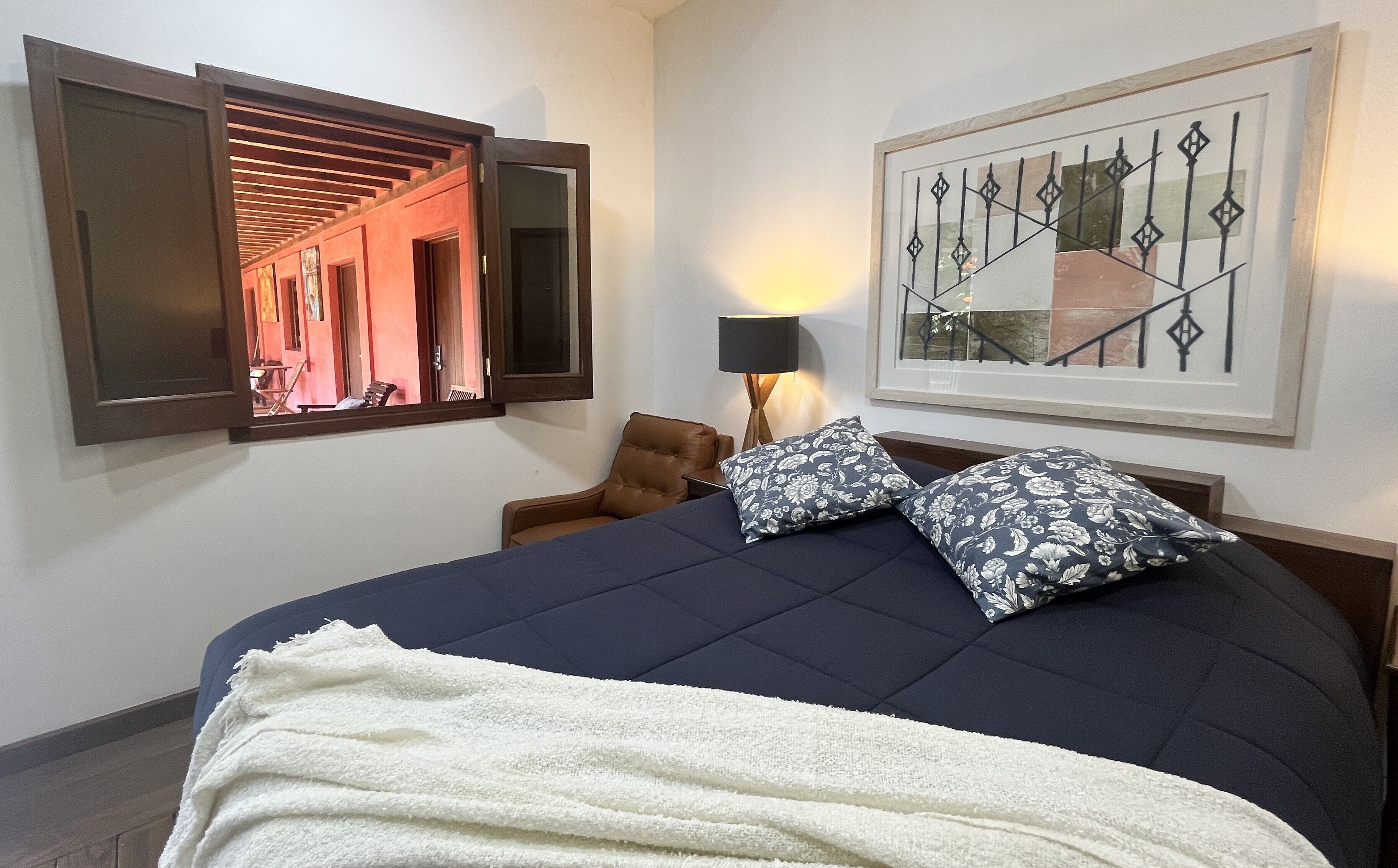 Une chambre d’hôtel avec un lit, une chaise, une lampe, un tableau encadré et une fenêtre donnant sur un couloir.
