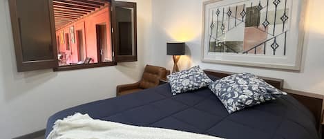 Une chambre d’hôtel avec un lit, une chaise, une lampe, un tableau encadré et une fenêtre donnant sur un couloir.