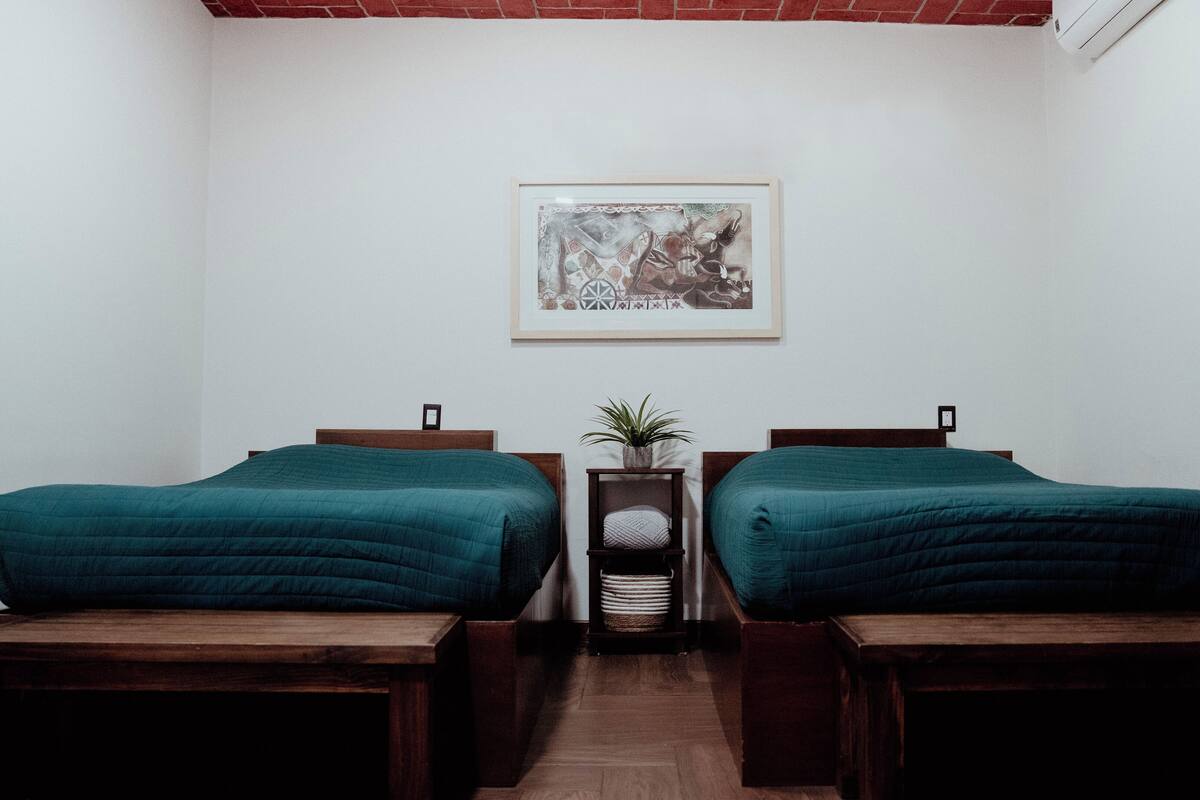 Ein Zimmer mit zwei Betten, einem kleinen Tisch mit einer Pflanze und einem gerahmten Bild an der Wand.