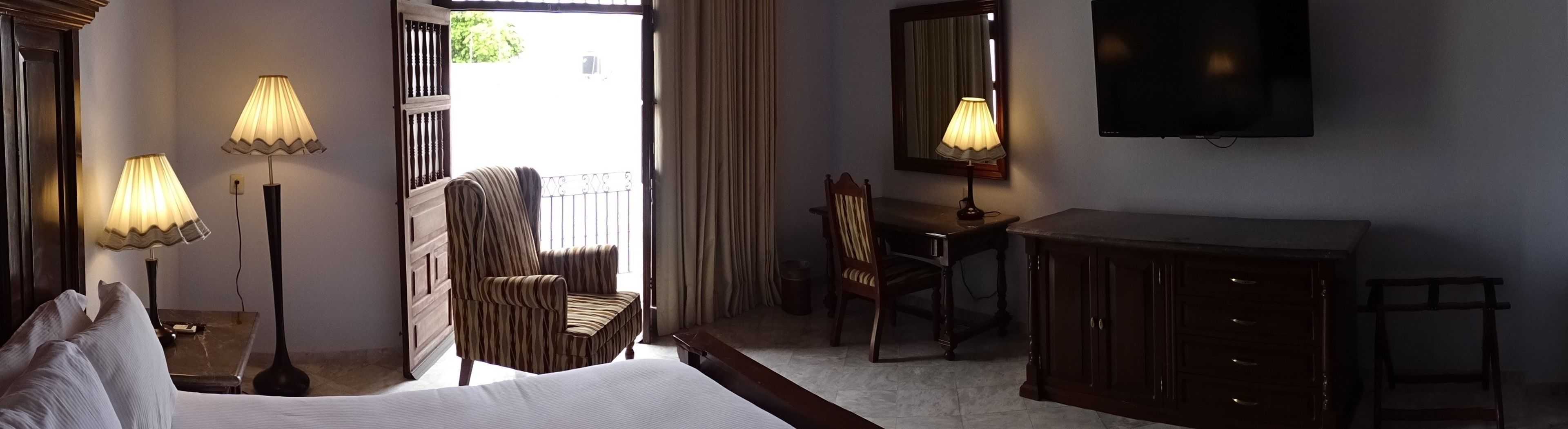 Quarto de hotel com uma cama, duas luminárias de cabeceira, uma escrivaninha com cadeira, um guarda-roupa e uma televisão.