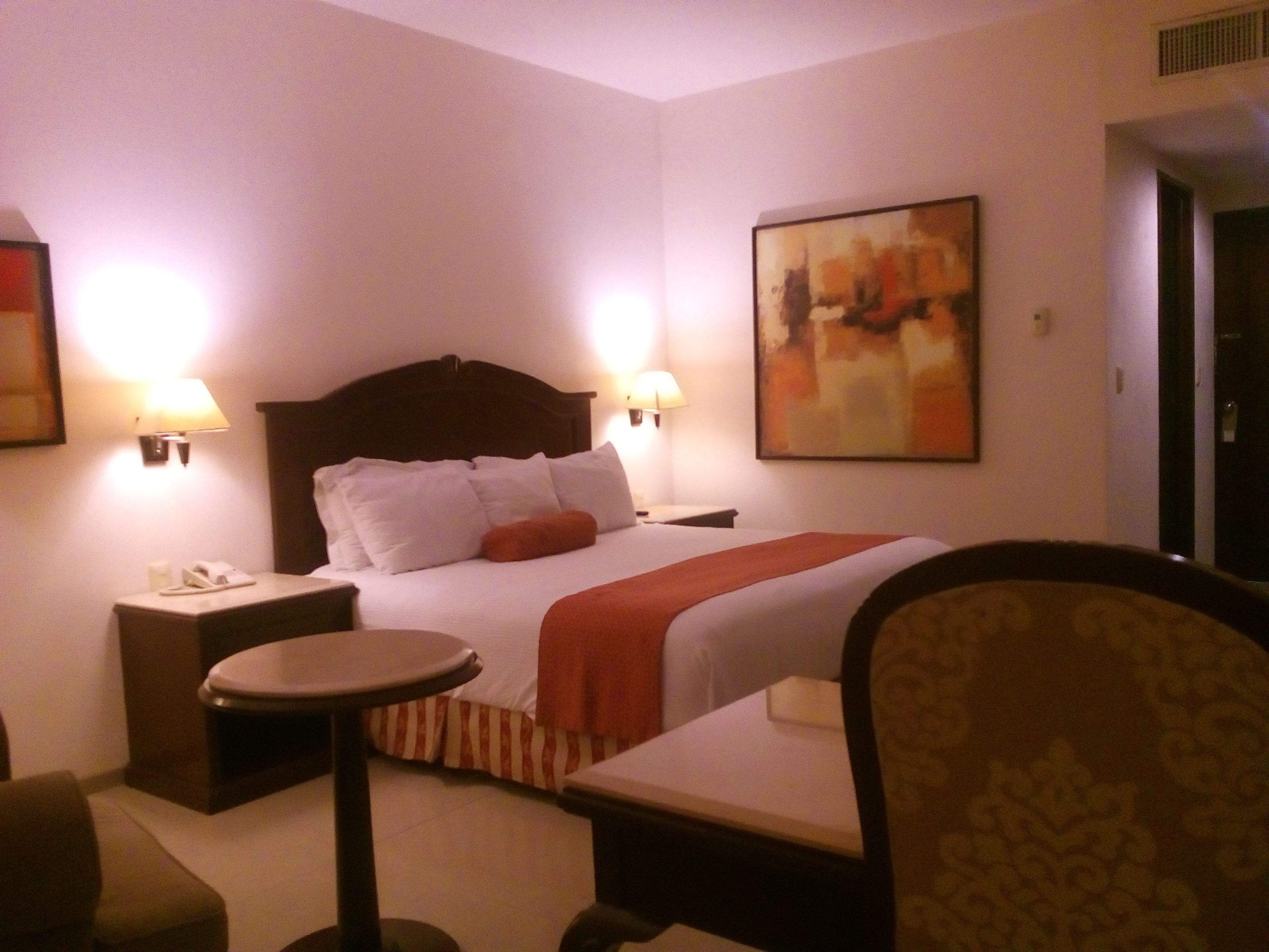 Una habitación de hotel con una cama, dos mesitas de noche, una silla y un cuadro en la pared.