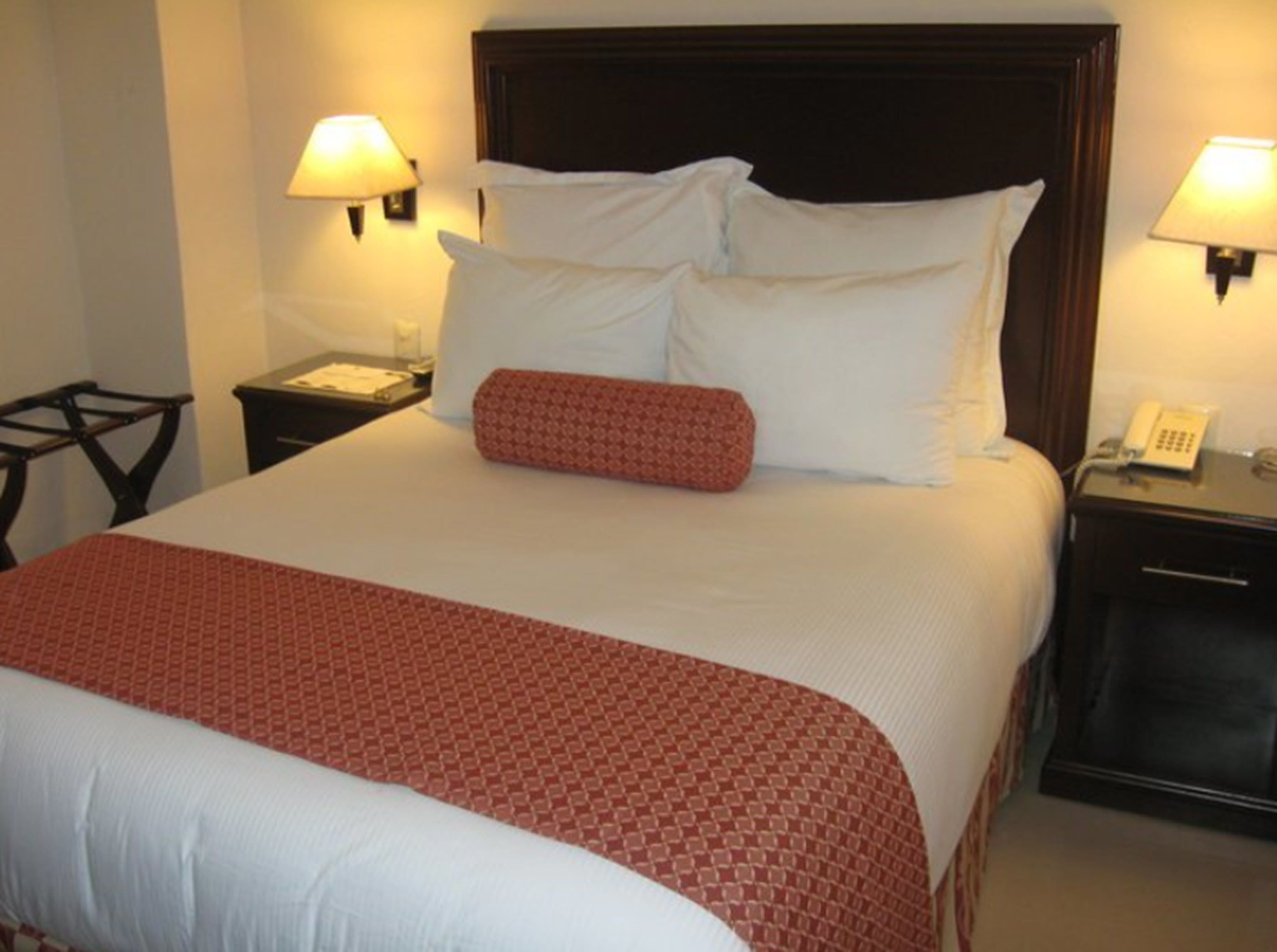 Una cama bien hecha con ropa de cama blanca y roja, dos mesitas de noche con lámparas y un teléfono en una de ellas.