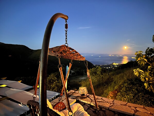 A swing chair with a woven seat and metal frame, overlooking a coastal landscape with a view of the sea and distant city lights. - Sokraki Villas (Corfu)