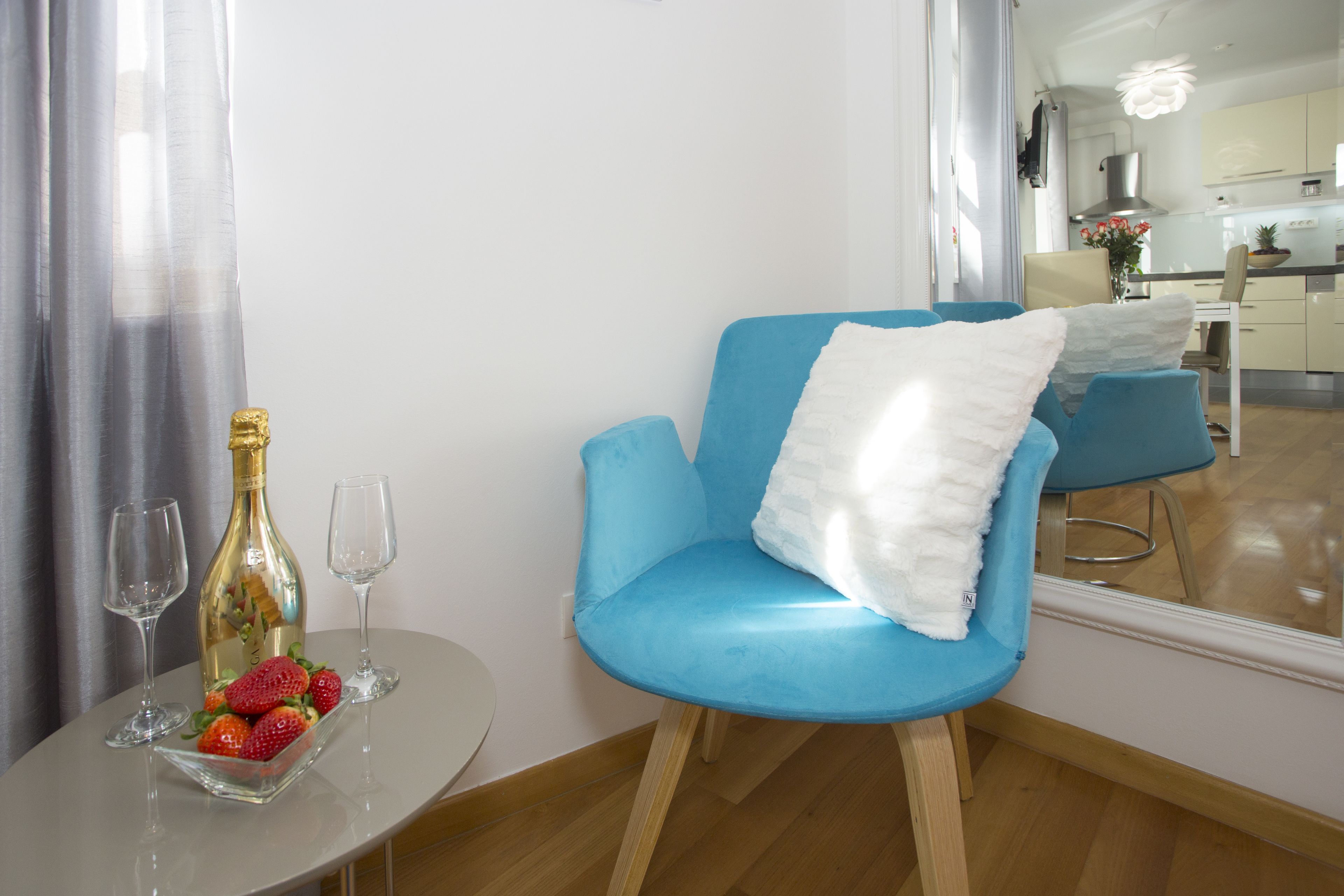 Ein modernes Zimmer mit einem blauen Sessel, einem kleinen Tisch mit einer Flasche und Erdbeeren sowie Blick auf eine Küche und einen Essbereich.