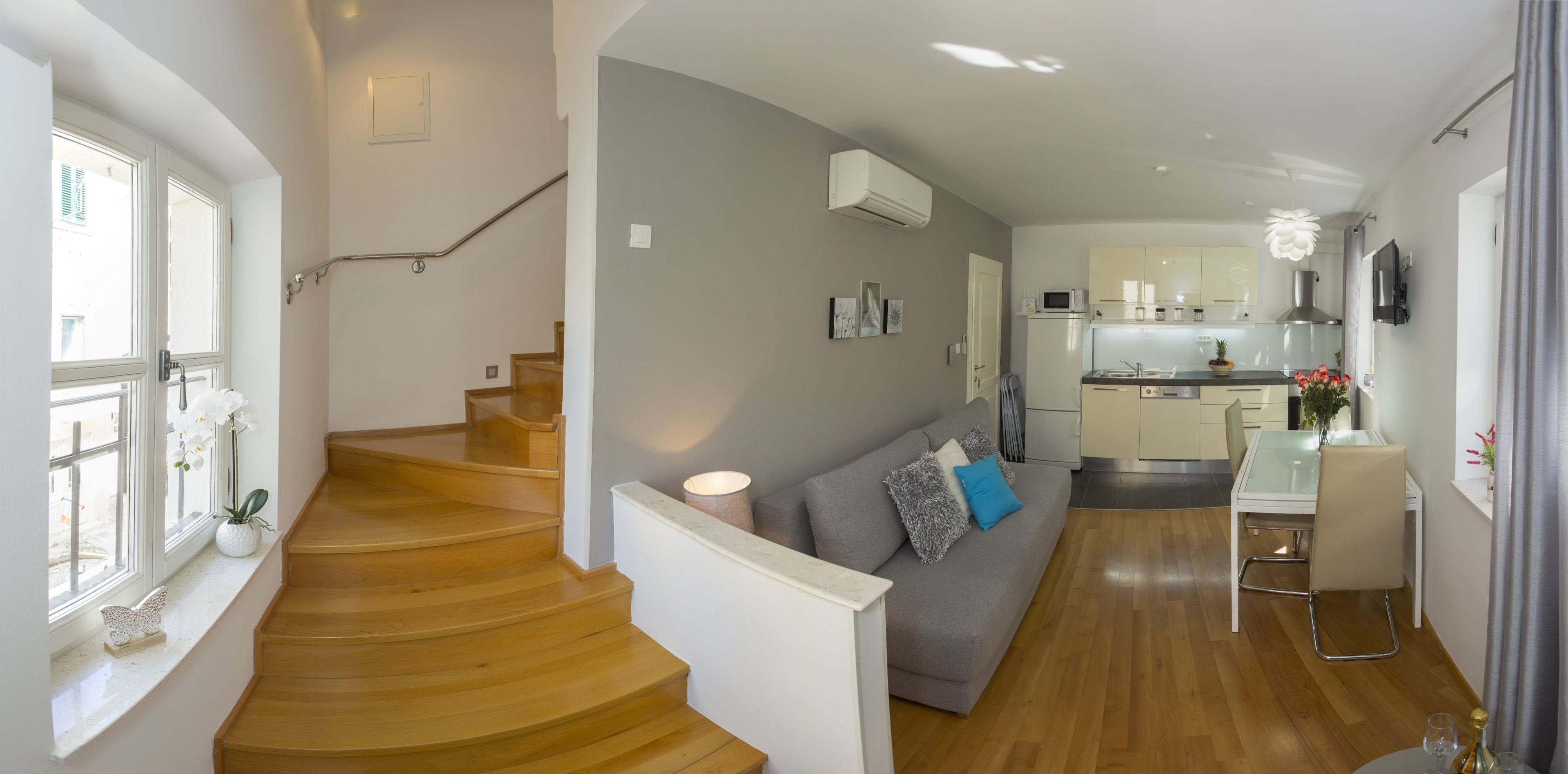 A modern apartment with a wooden staircase, a grey sofa with blue cushions, a dining area with a table and chairs, and a kitchen with white cabinets and a microwave.
