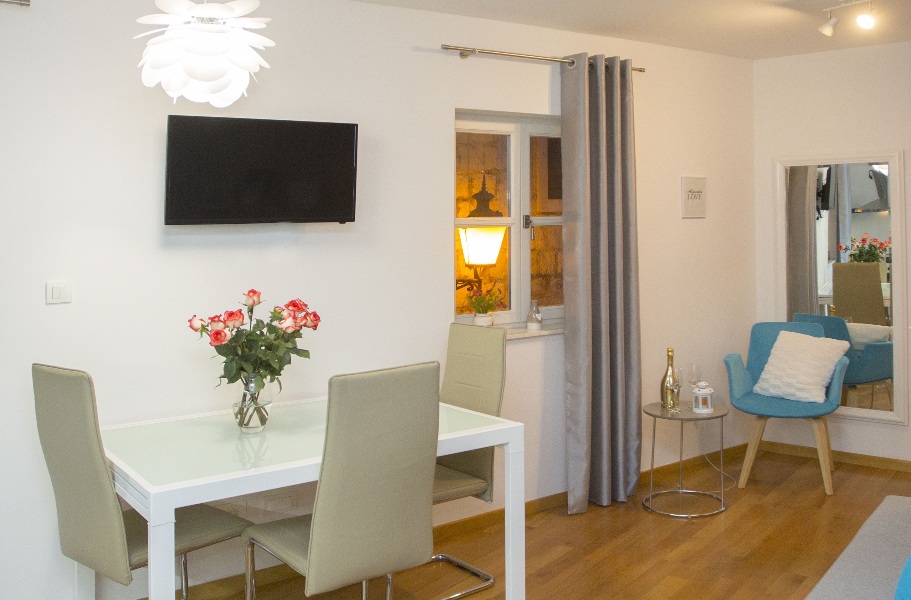 A modern living room with a flat-screen TV, a dining table with chairs, a vase of flowers, a blue armchair, and a small side table with a bottle and candle.