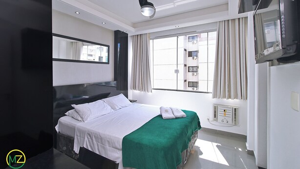 Habitación de hotel moderna con una cama grande, una manta verde, un televisor de pantalla plana y un amplio ventanal con cortinas.
