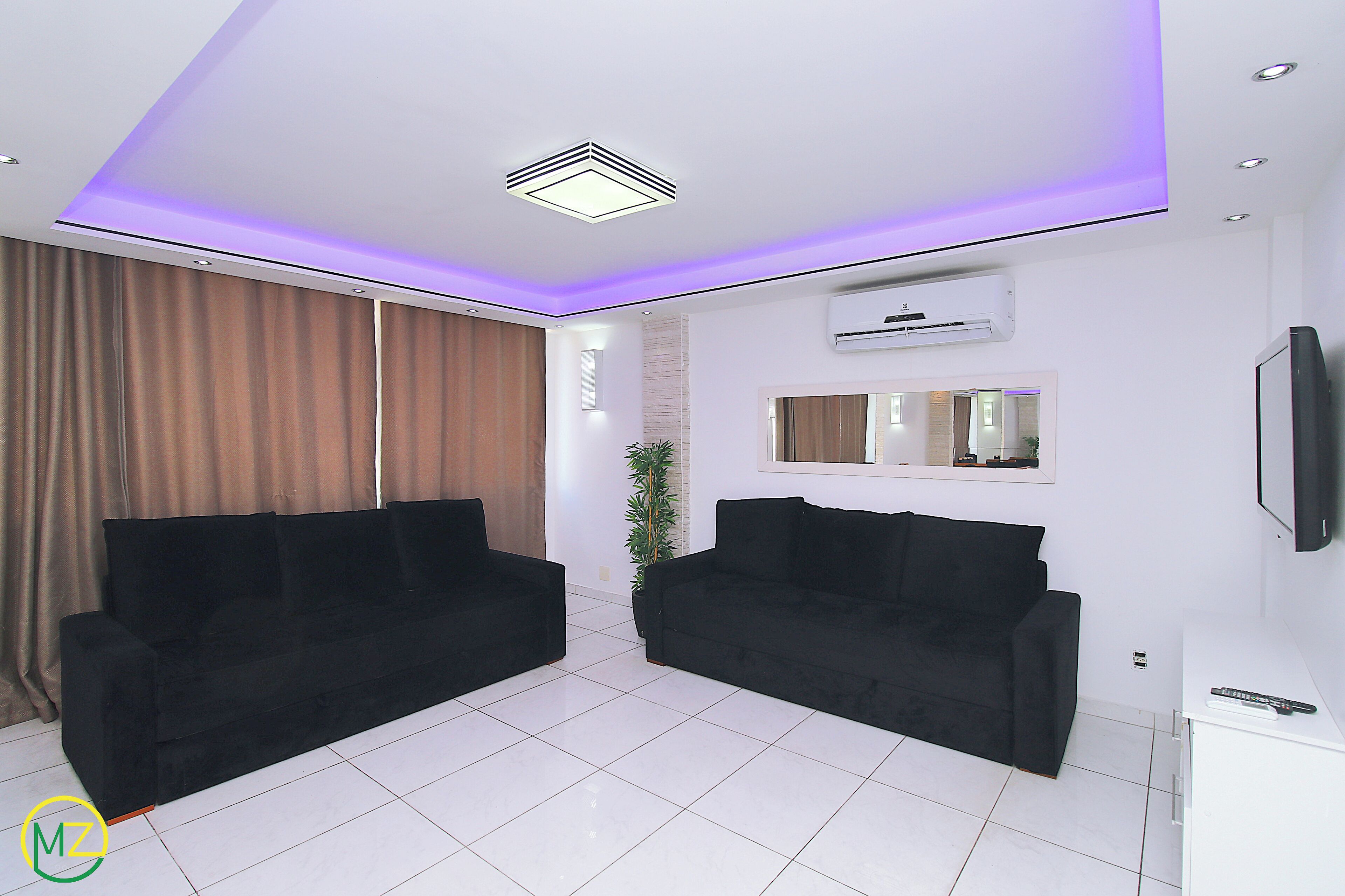 A modern living room with two black sofas, a white tiled floor, a ceiling with recessed lighting, an air conditioning unit, and a flat-screen TV mounted on the wall.