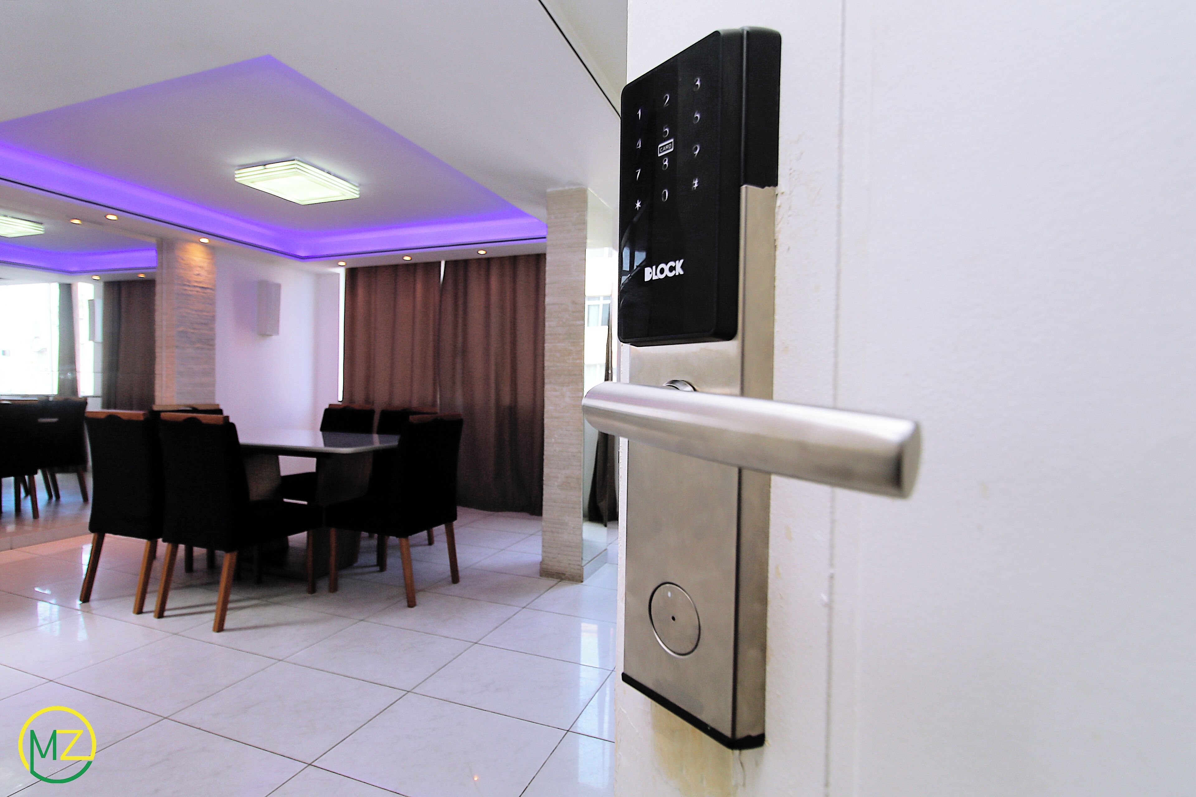 A modern hotel room with a digital lock on the door, a dining area with a table and chairs, and a ceiling with recessed lighting.