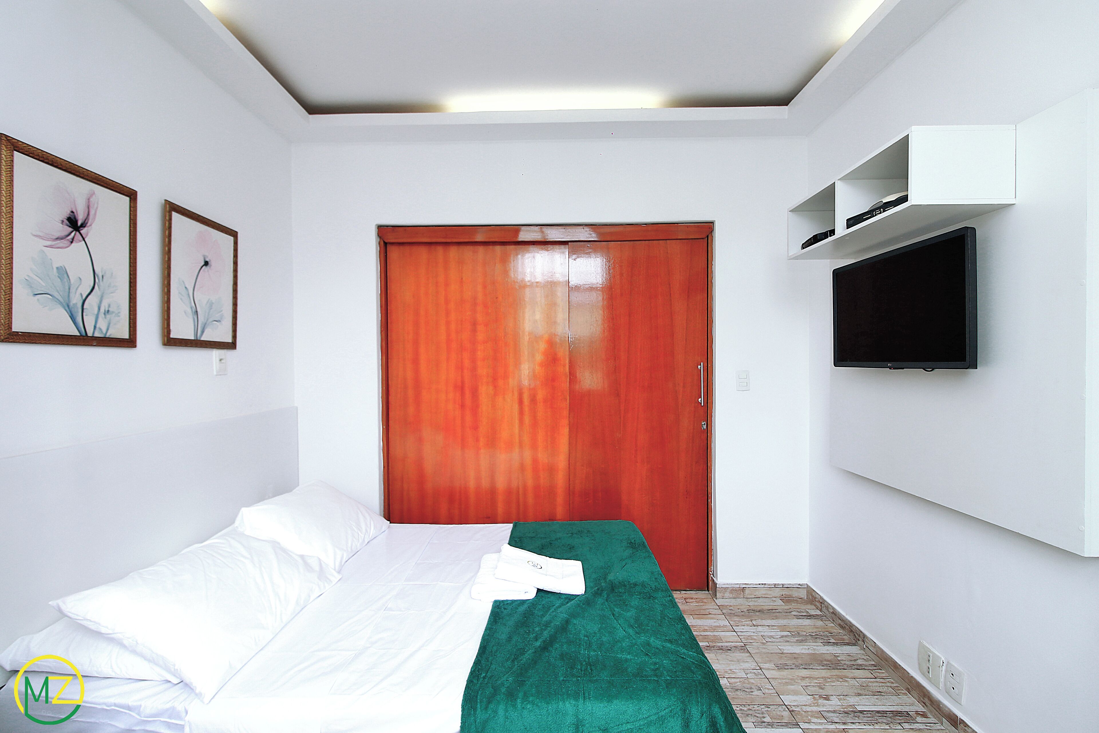 Habitació d'hotel amb un llit, una manta verda, un armari de fusta, un televisor fixat a la paret i dues obres florals emmarcades.
