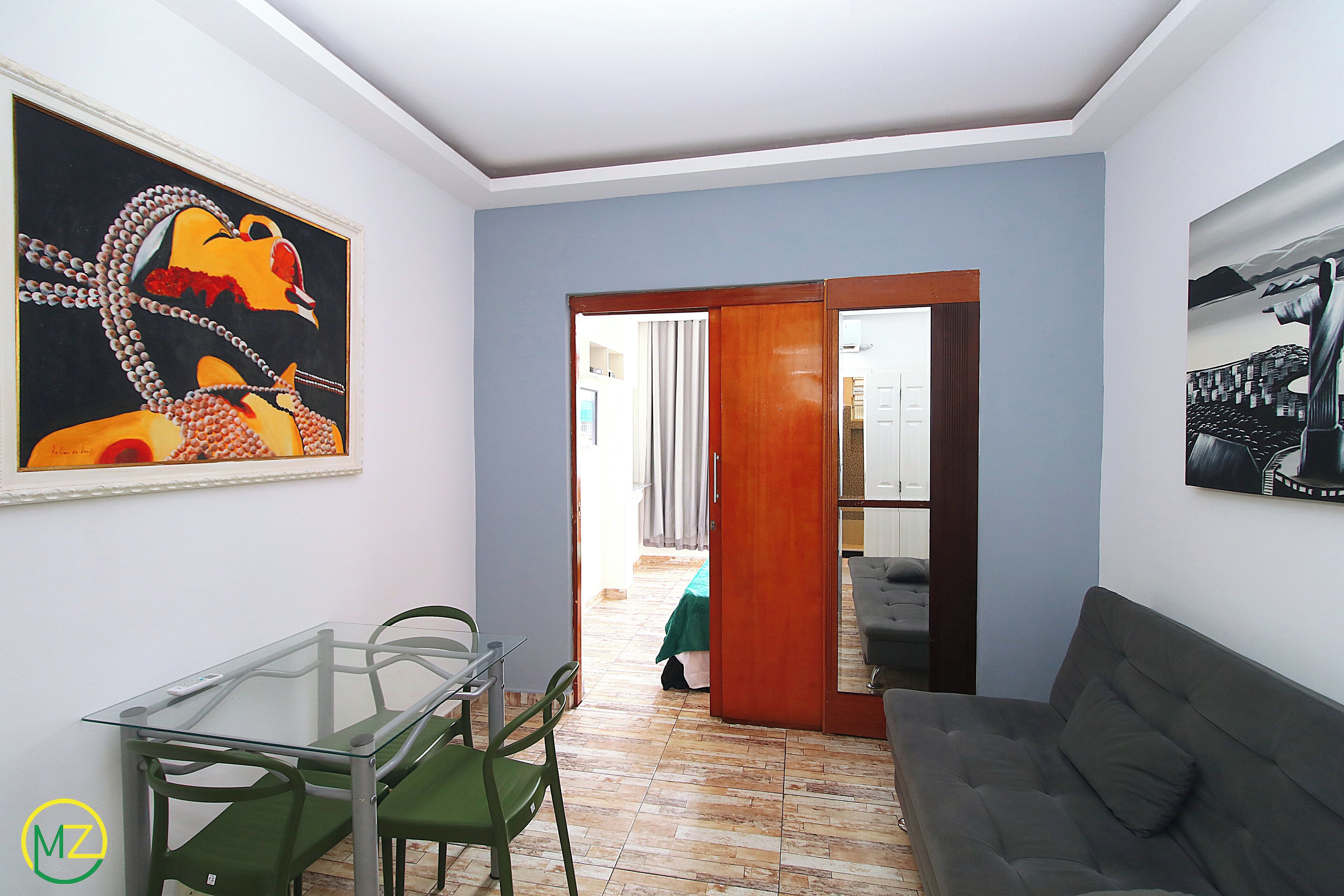 A room with a glass table, green chairs, a grey sofa, and two paintings on the walls.