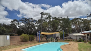 Une piscine rectangulaire à bords incurvés, entourée d’une terrasse en briques. Un espace ombragé est aménagé avec un auvent jaune, une table et des chaises.