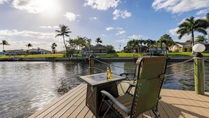 Une terrasse en bois avec une table et un fauteuil roulant, donnant sur une étendue d’eau calme, avec des palmiers et des maisons en arrière-plan.
