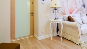 Een kamer met een glazen deur, een witte lamp op een klein tafeltje, een witte bank en een grote bloemenmuur.