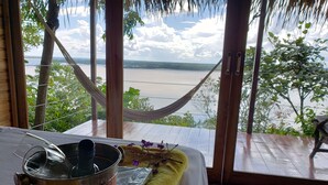 A room with a view of a river, a hammock, a table with a pot and a glass, and a wooden floor. - Alta Vista Amazon Lodge (Manacapuru)