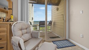 A room with a sliding glass door leading to a balcony, a cushioned chair, a small table, and a wooden dresser. - Beachfront and Gorgeous Sunrise Views - A Beach Lover's Paradise  (South Padre Island)