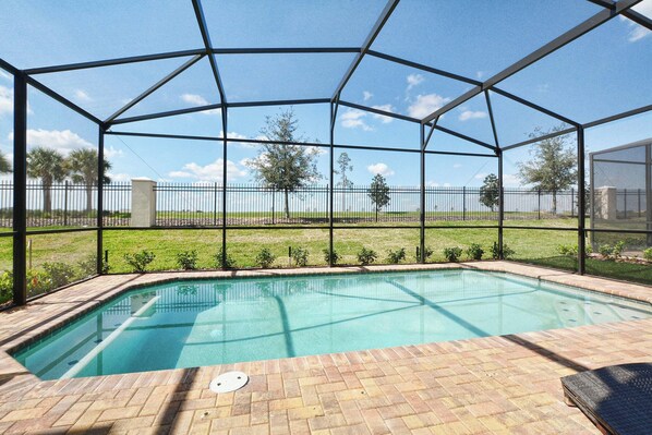 A covered outdoor swimming pool with a brick patio, surrounded by a metal fence and trees in the background.