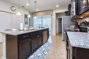 A kitchen with dark cabinetry, granite countertops, a central island, pendant lighting, and a view into the dining area. - 5BR Windsor Pool Home | 3 Master Suites | Game Room | Dog Friendly (Kissimmee)