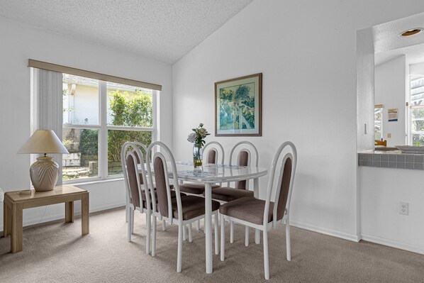 Un coin repas avec une table ronde et six chaises, une petite table d’appoint avec une lampe, un tableau encadré au mur et une fenêtre donnant sur un paysage verdoyant.