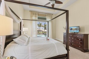 Een hemelbed met witte lakens, een televisie op een dressoir, en uitzicht op een zwembad en palmbomen door het raam.