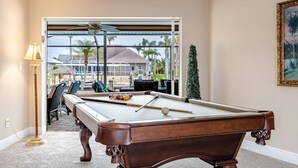 A pool table with balls, a ceiling fan, and a view of outdoor seating and palm trees. - Boater's Delight | Gulf Access, Bikes, Kayaks, Pool Table, Heated Poo/Spa - Villa Blue Sky - Roelens (Cape Coral)