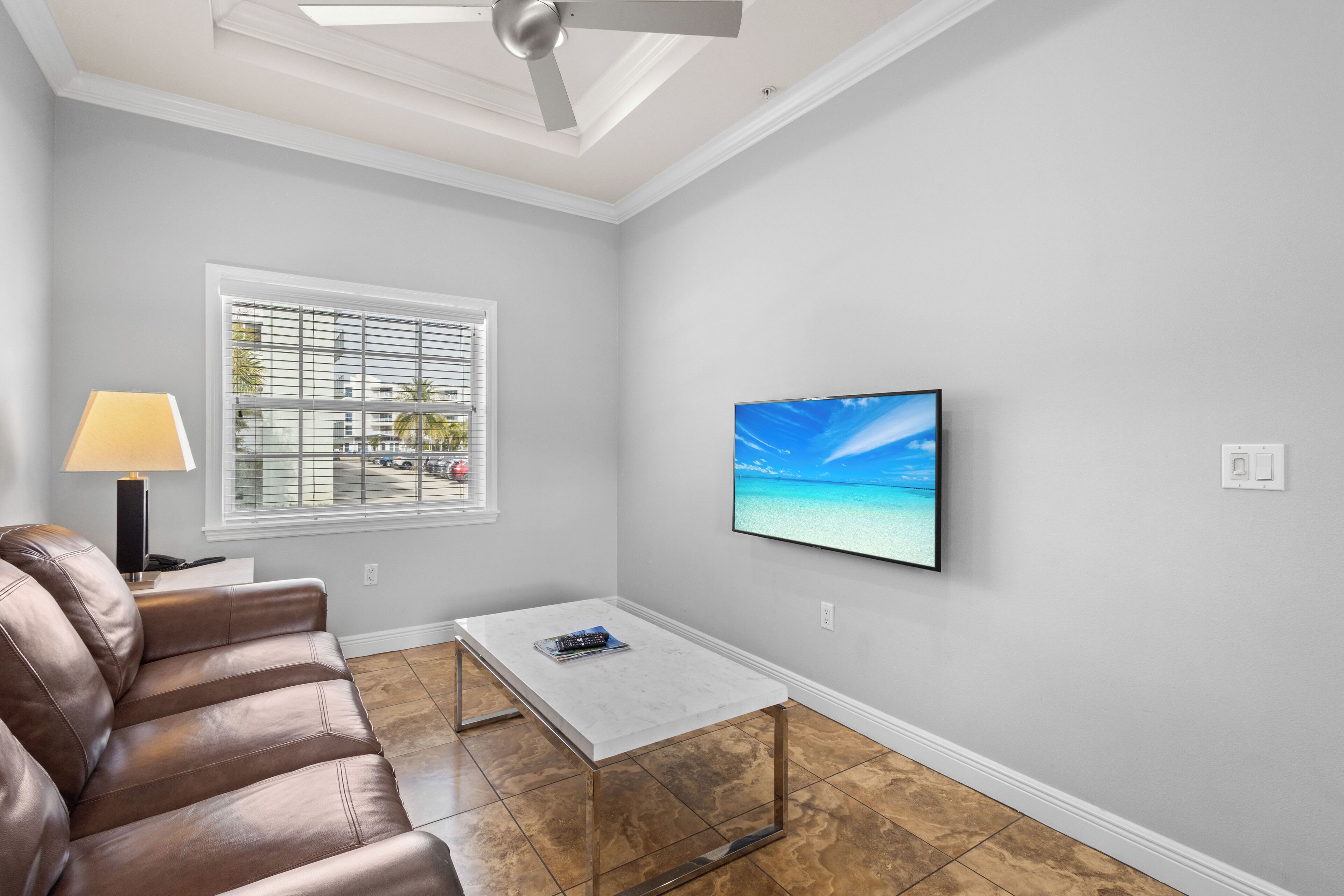 A living room with a leather sofa, a coffee table, a TV mounted on the wall, and a window with blinds.