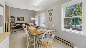 A dining area with a wooden table and white chairs, a kitchen area with a refrigerator and cabinets, and a window with a view of trees.