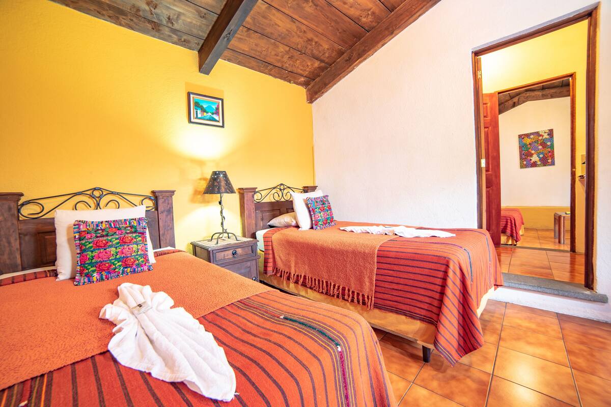 Una habitación de hotel con dos camas, cabecero de madera, una mesita de noche, una lámpara y un cuadro en la pared.