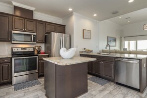 A modern kitchen with stainless steel appliances, a marble island, and dark wooden cabinets. - Ocean Melody | Elevated Beach Living (Dauphin Island)