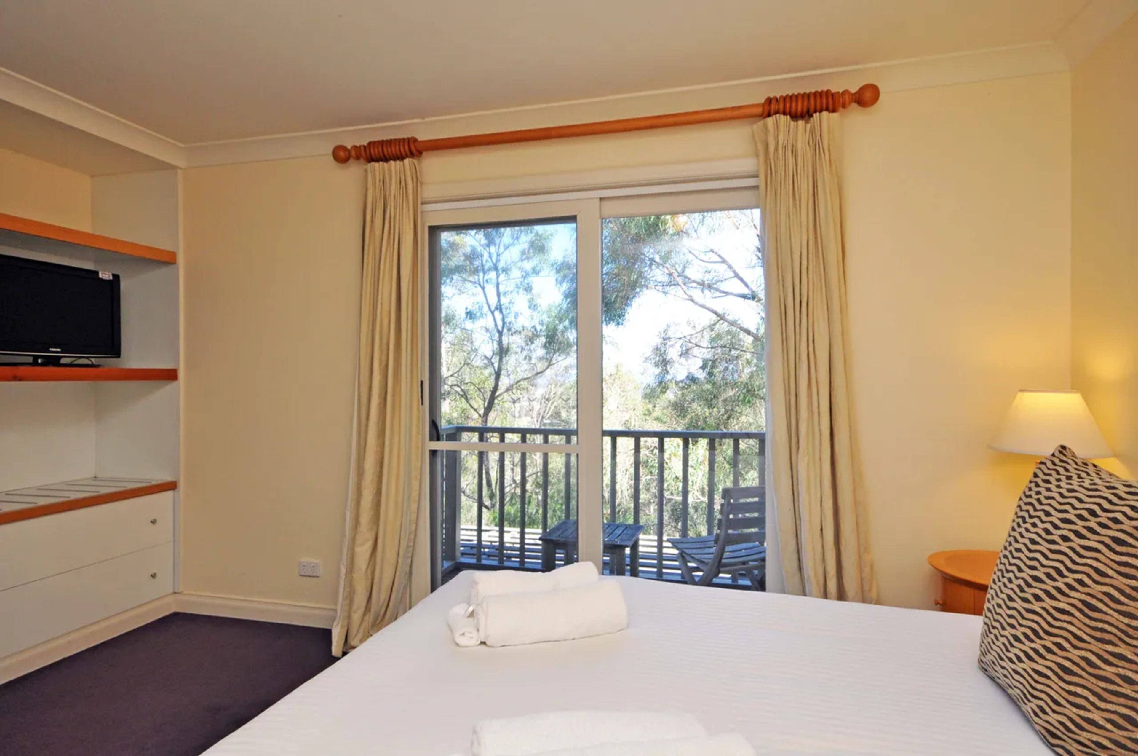 Une chambre d’hôtel avec un lit, une télévision, un balcon avec des chaises et une fenêtre avec des rideaux.